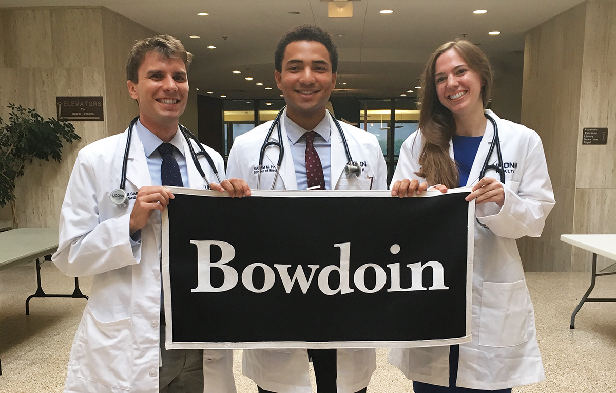 3名白色医疗外套的学生举起Bowdoin标志 